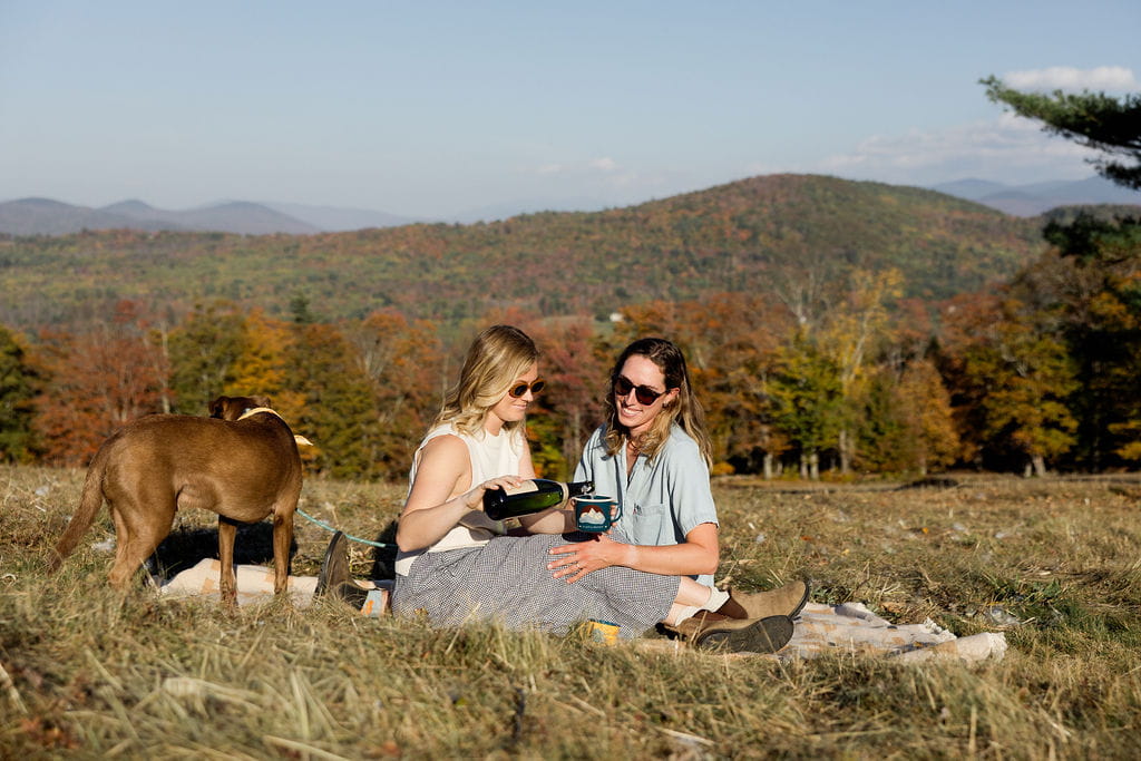 Two women picnic on a rolling hill with their dog during their adventure couples session.