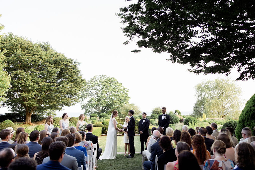 Guests sit in white wood chairs during an Eolia Mansion wedding ceremony in the garden.